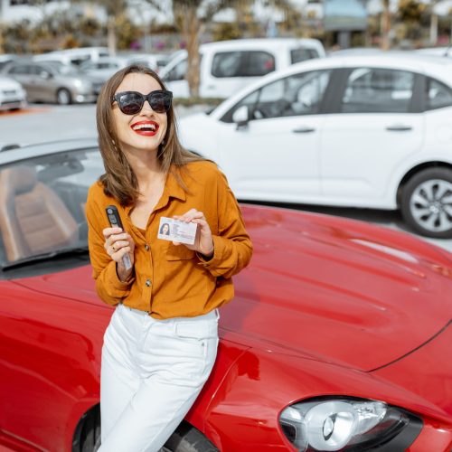 Portrait of a beautiful young woman standing with driver's license and keys near the red cabriolet at the car parking. Concept of a happy buying or renting a car